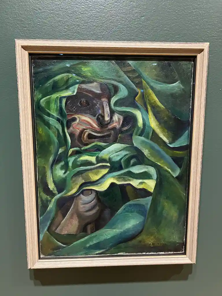 A framed painting by Emily Carr, depicting a distorted, expressive face partially obscured by swirling green and yellow…