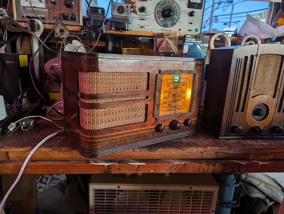 A vintage wooden RCA Victor radio with a glowing orange dial and woven speaker grille sits on a cluttered workbench…