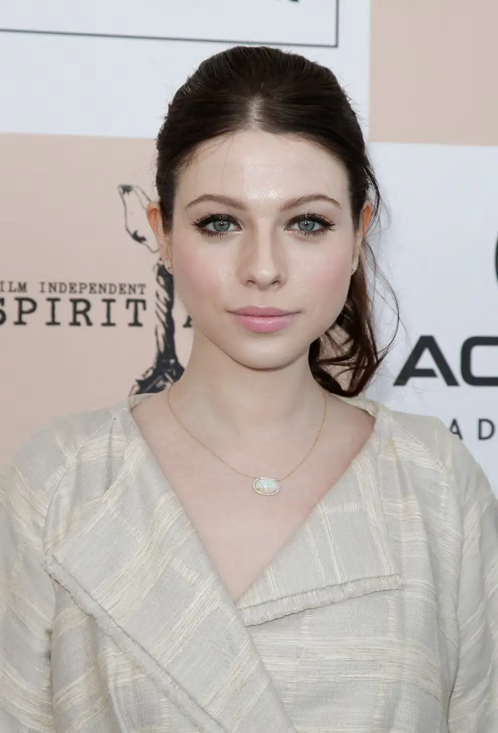 A woman with dark hair pulled back, wearing a light-colored textured top and a gold necklace with a white stone, looks…