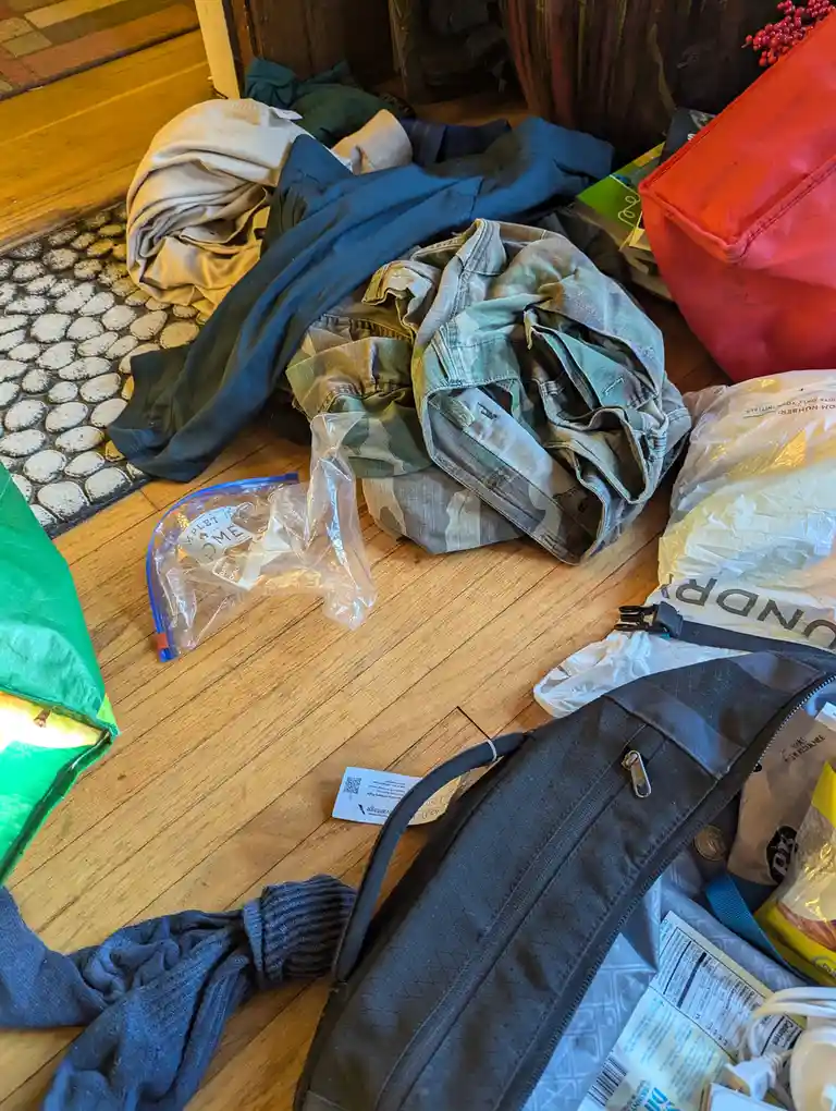 A messy floor is covered with clothes, bags, and various items like a plastic bag, a green tarp, and a black bag with…