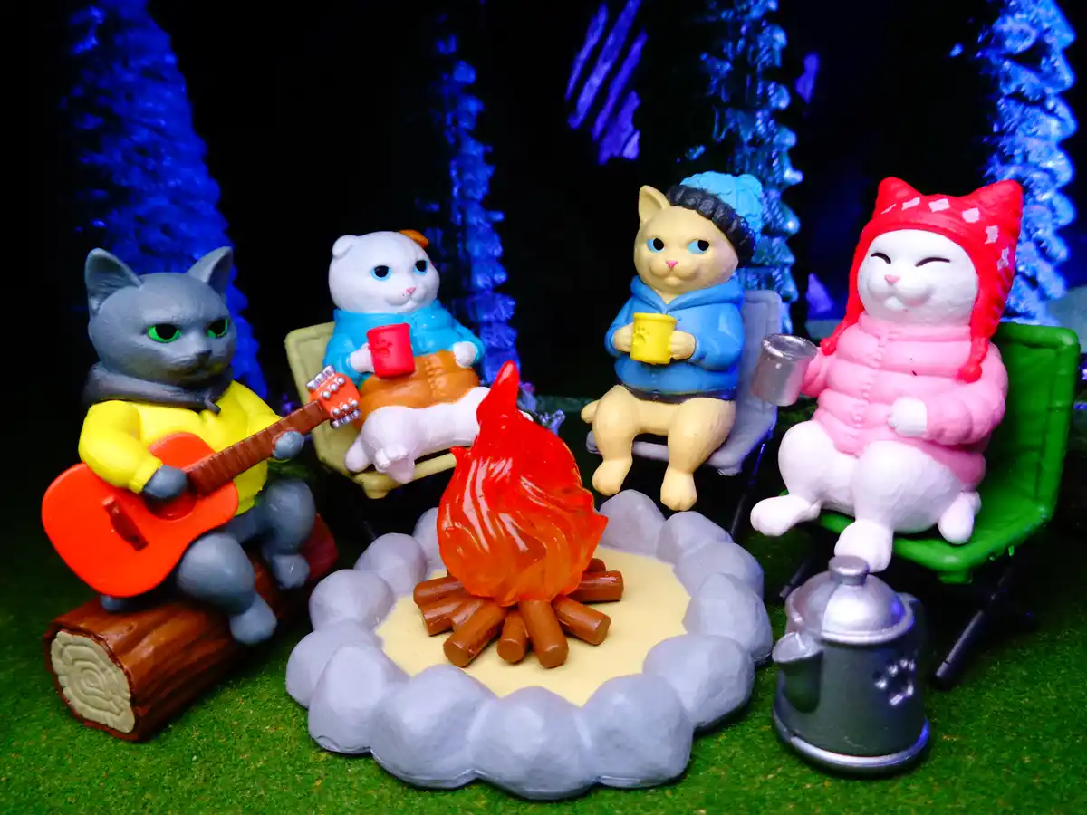 Four plastic cat figurines are gathered around a miniature campfire in a forest setting, each dressed in cozy winter…