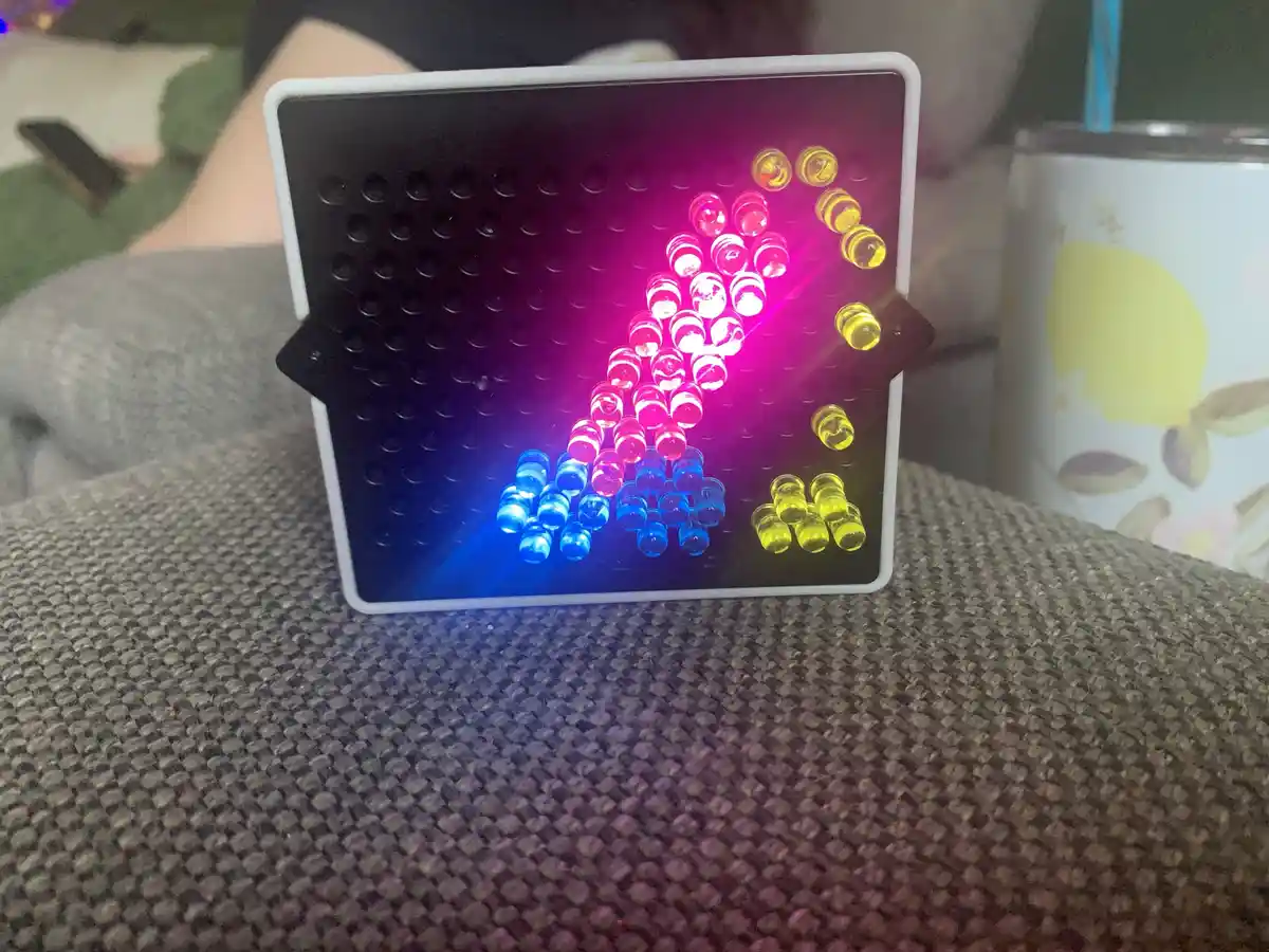 A small, square LED matrix display with a white frame sits on a textured gray couch, showing a pixelated image of a person…