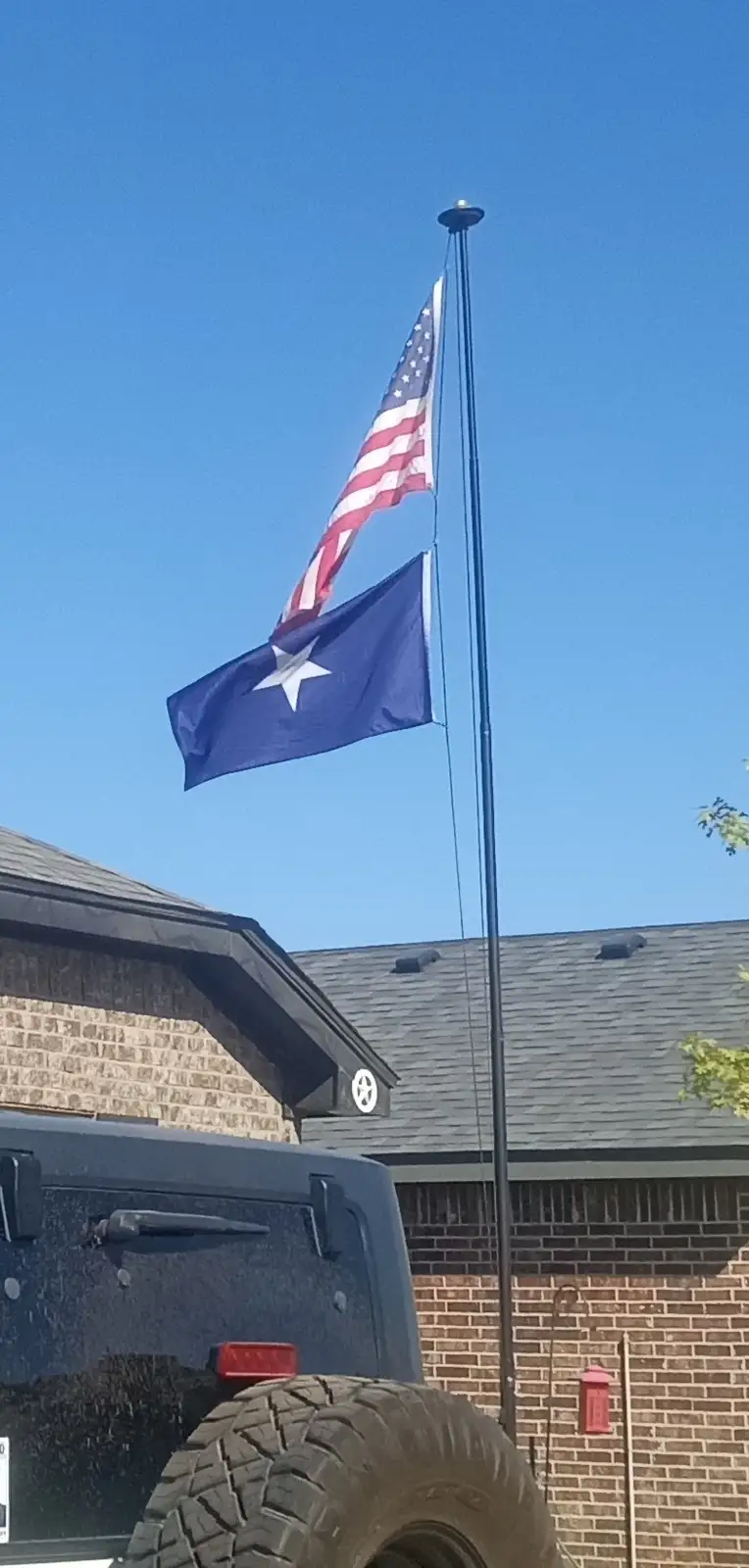 An american flag and a texas state flag are flying side-by-side on a single flagpole against a clear blue sky, with a brick…