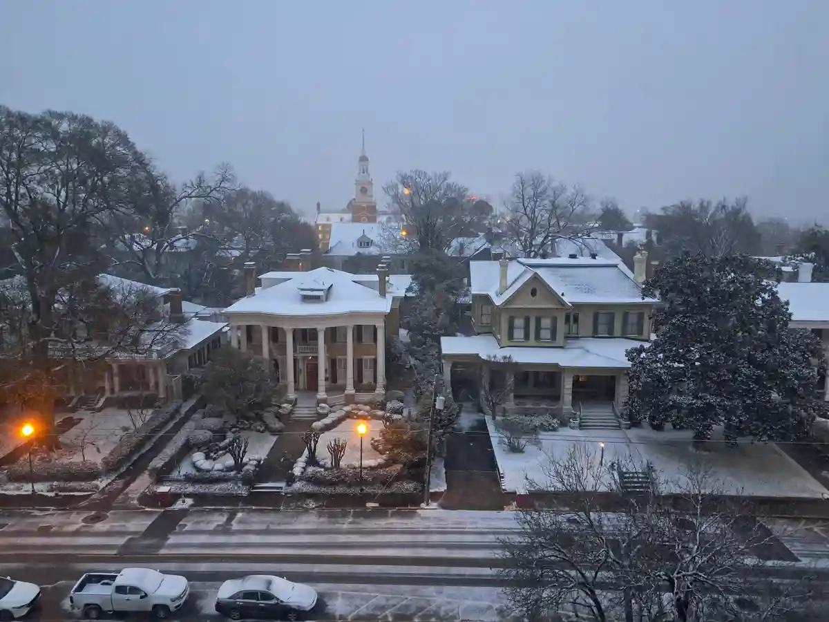 A snowy urban scene shows large, ornate houses with snow-covered roofs and yards, bare trees, and streetlights glowing…