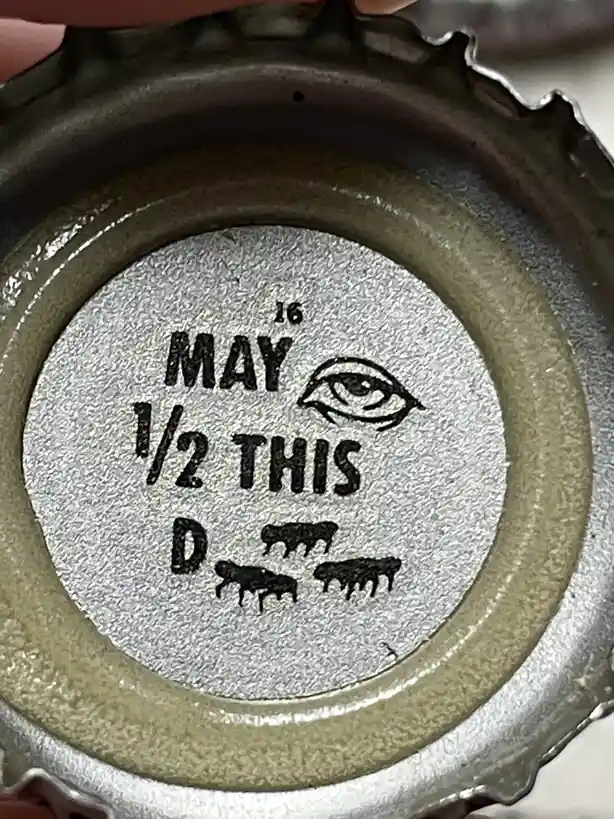 A close-up of the bottom of a bottle cap showing a stamped date '16 MAY' followed by '1/2 THIS D', with a drawn eye above…