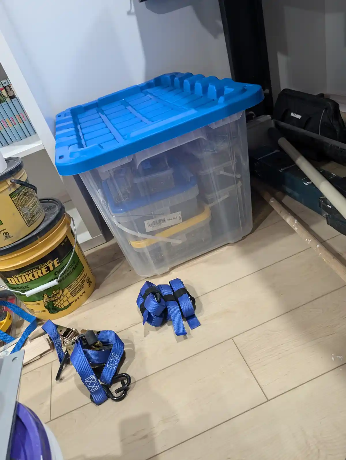 A cluttered garage floor shows blue ratchet straps, a large yellow Quikrete bucket, and a clear plastic storage bin with a…