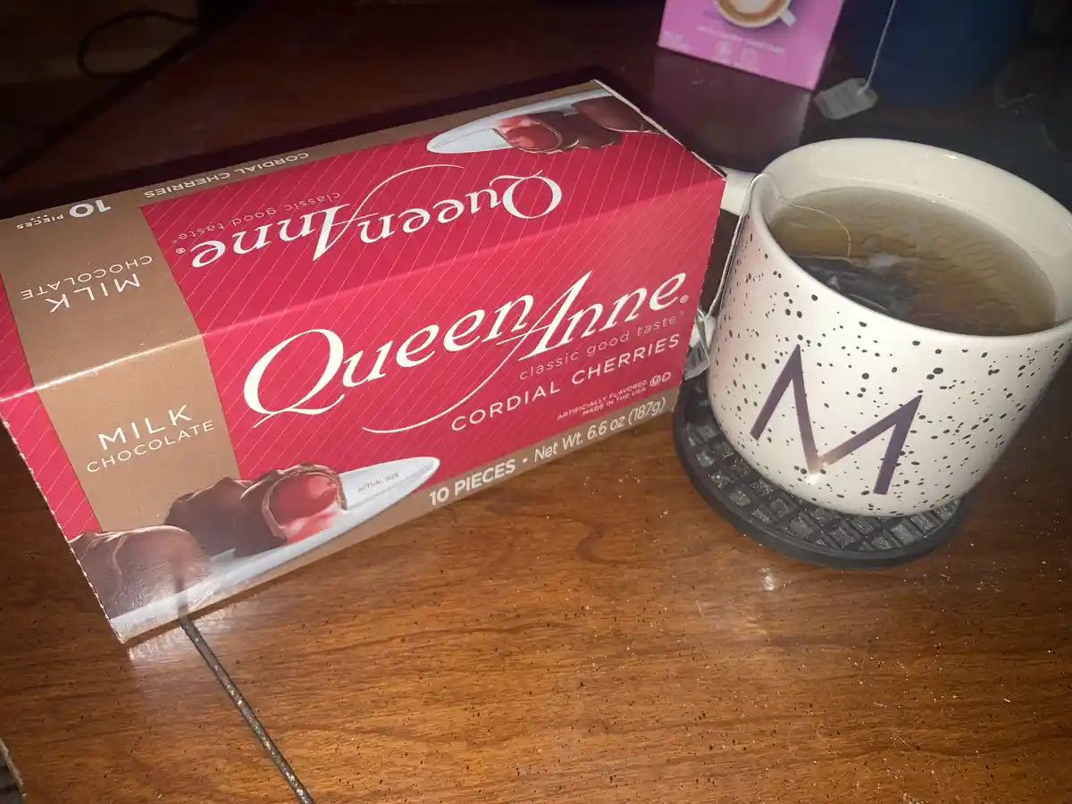 A red and brown box of Queen Anne milk chocolate cordial cherries sits next to a white speckled mug with a large purple 'M'…