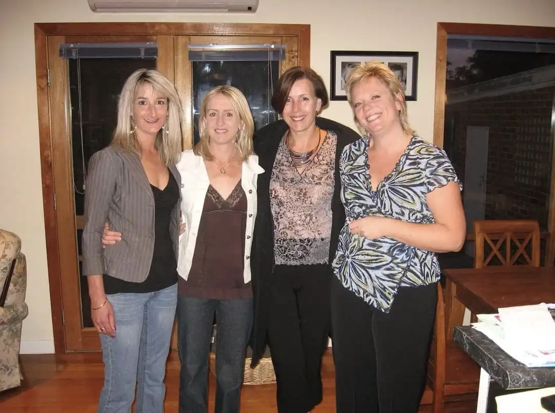 four smiling women posing together indoors, arms around each other, in a casual home setting with wooden doors and furniture…