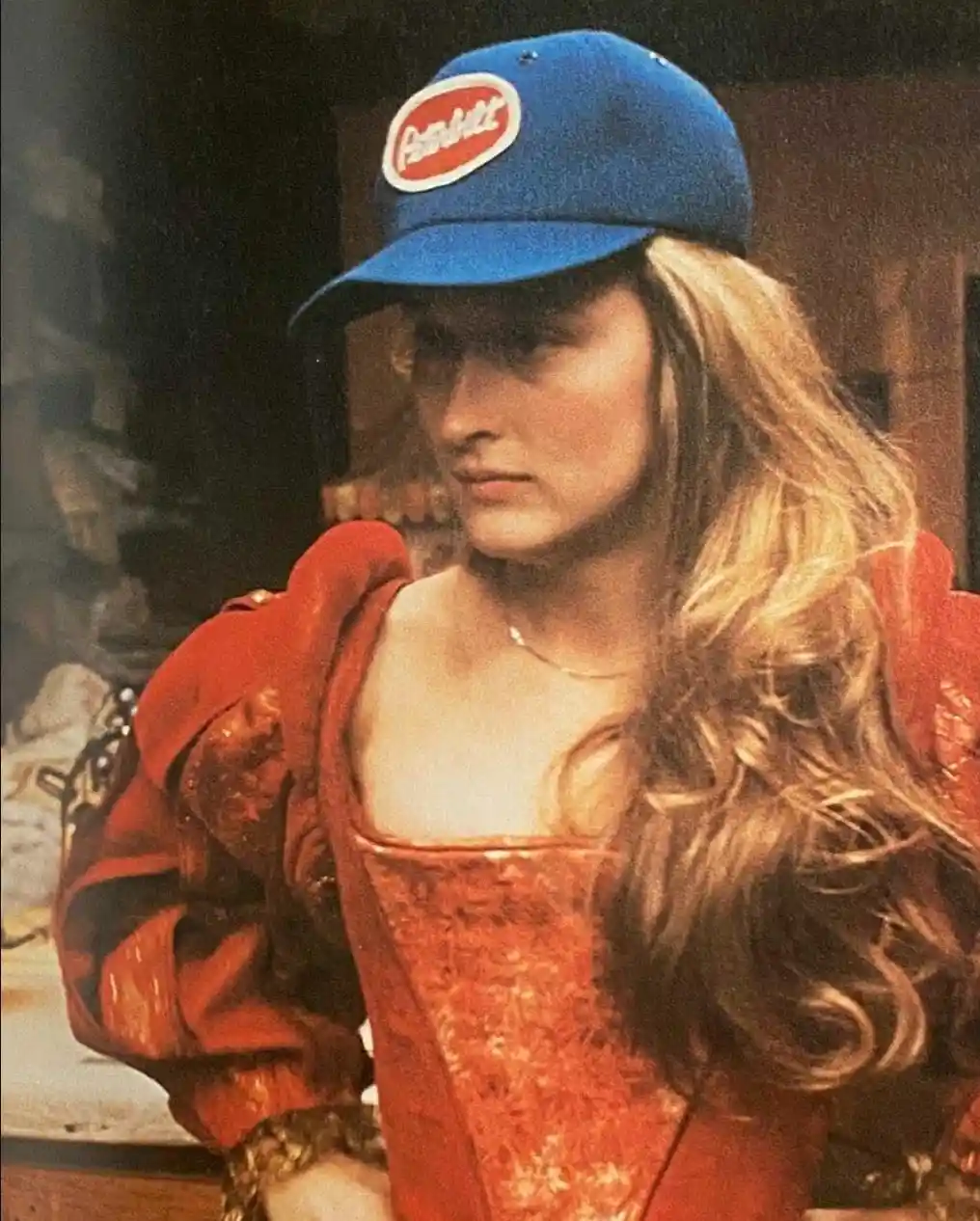 a woman with long blonde hair wearing a red, ornate, historical-style dress and a blue baseball cap with a red oval logo…