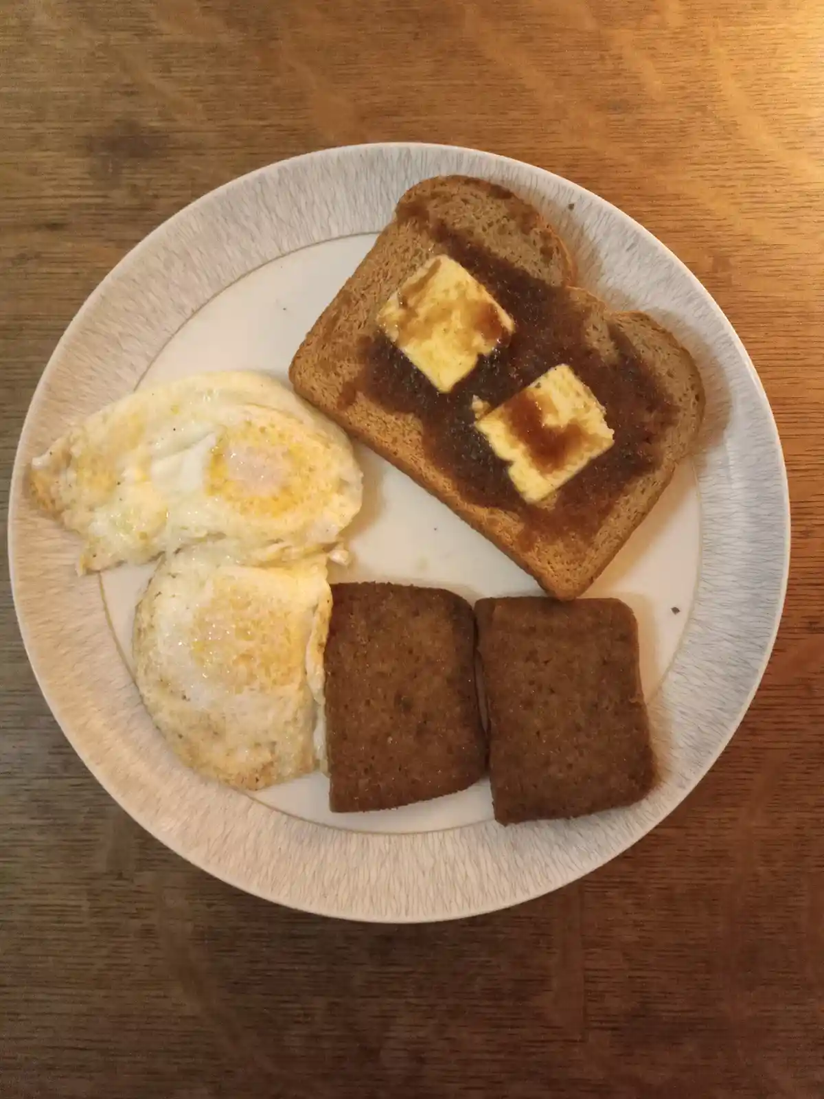 A plate of breakfast food including two fried eggs, a slice of toast with butter and syrup, and two small brown squares that…