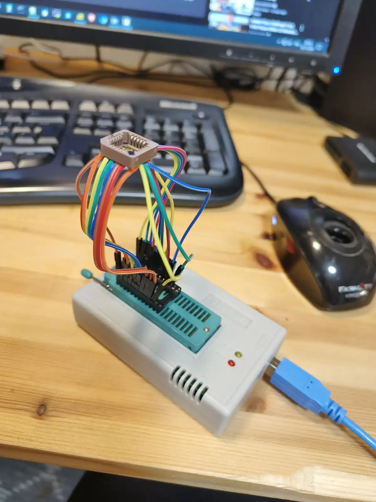 A close-up photo shows a homemade electronic device on a wooden desk, featuring a white USB programmer with a teal…