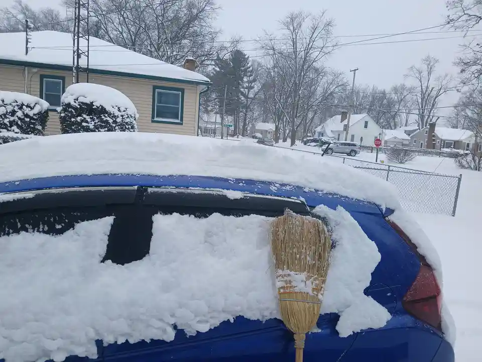 A blue car is heavily covered in snow, with a broom stuck upright in the rear window area, partially buried in snow. The…