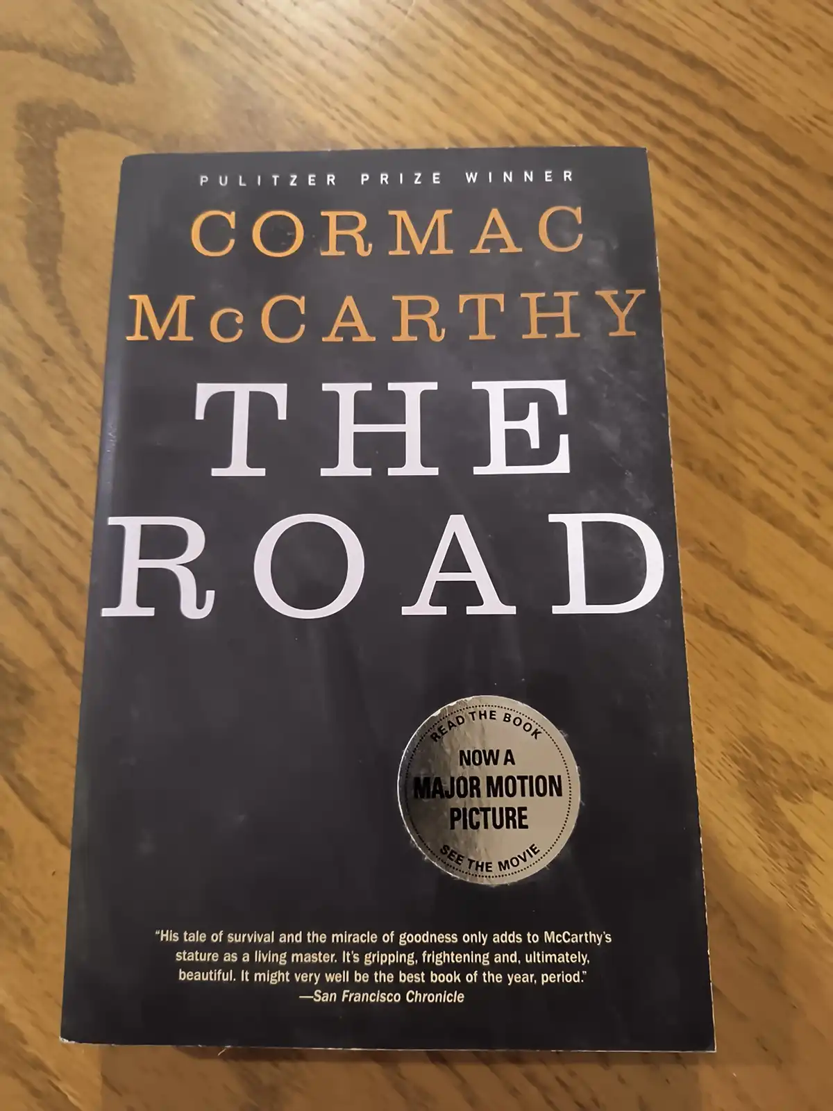 A physical copy of Cormac McCarthy's Pulitzer Prize-winning novel 'The Road' rests on a wooden surface. The book cover is…