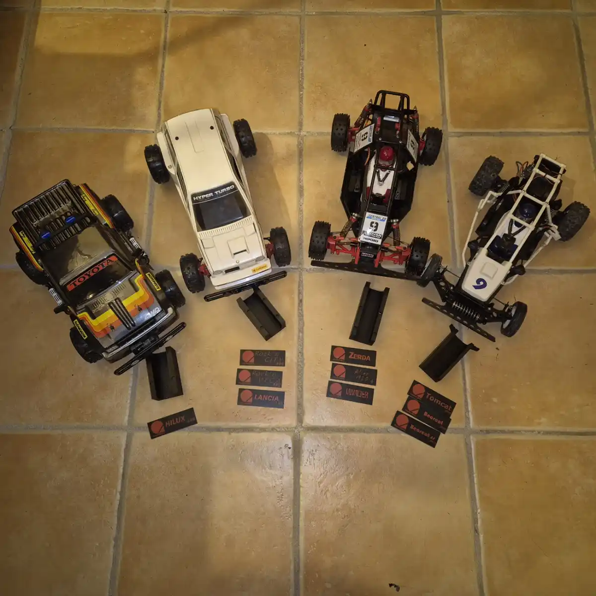 Four radio-controlled off-road vehicles are arranged on a tiled floor, each with a set of black and red decals placed…
