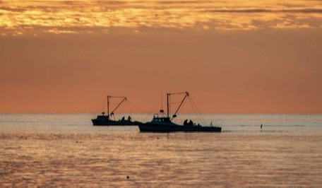 Two fishing boats are silhouetted against a vibrant orange and yellow sunset over calm ocean waters. The boats are…
