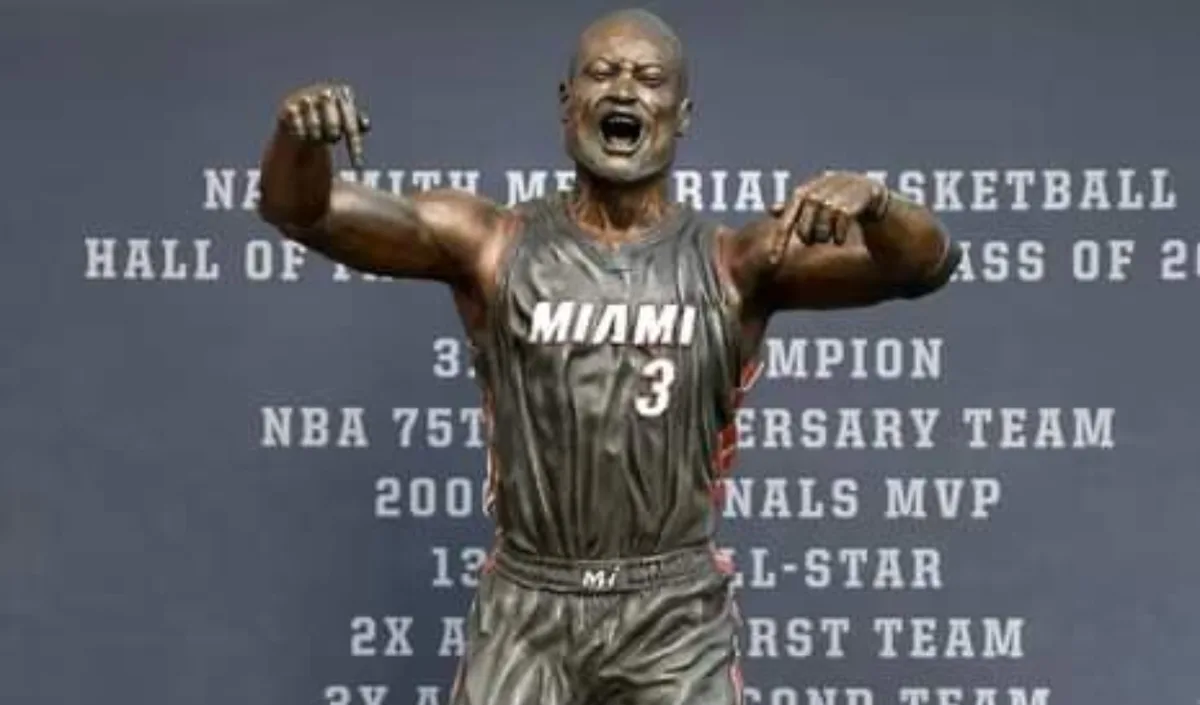 A bronze statue of a basketball player in a Miami jersey with number 3, arms raised and pointing down, stands before a…