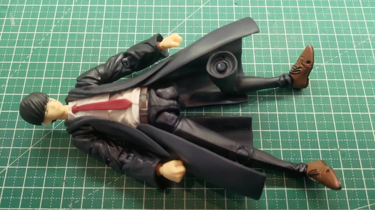 A plastic action figure of a man with black hair, wearing a black trench coat, white shirt, red tie, and brown shoes, lies…