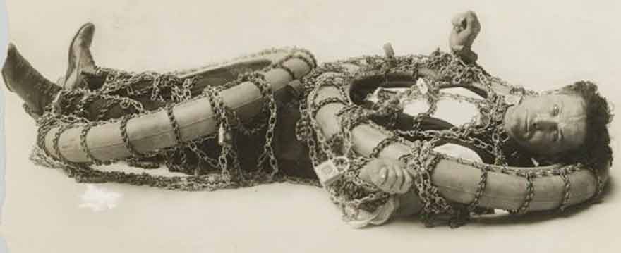 a sepia-toned photograph shows a man lying on his back, completely entangled in heavy chains that wrap around his torso…