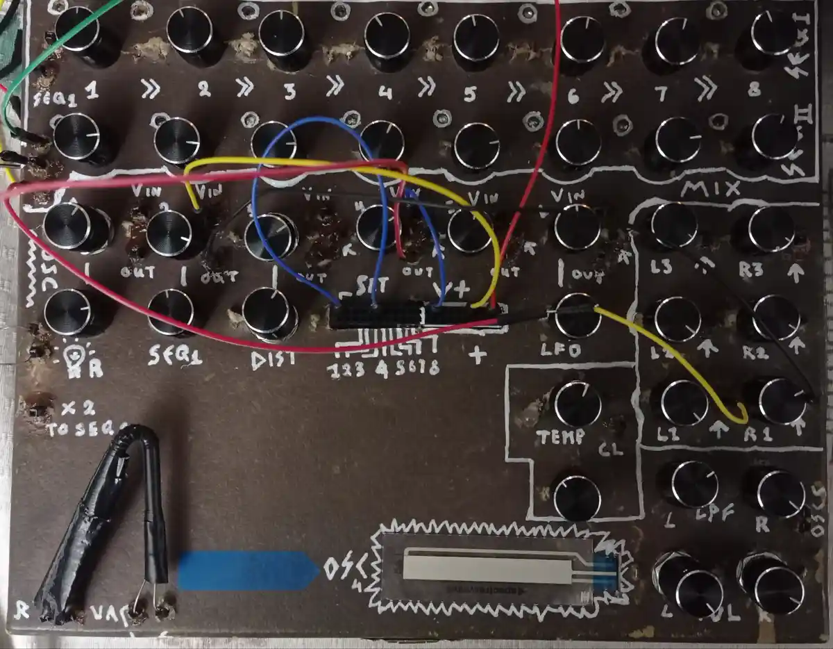 A close-up photo of a homemade electronic music synthesizer built on a dark brown breadboard, featuring numerous black…
