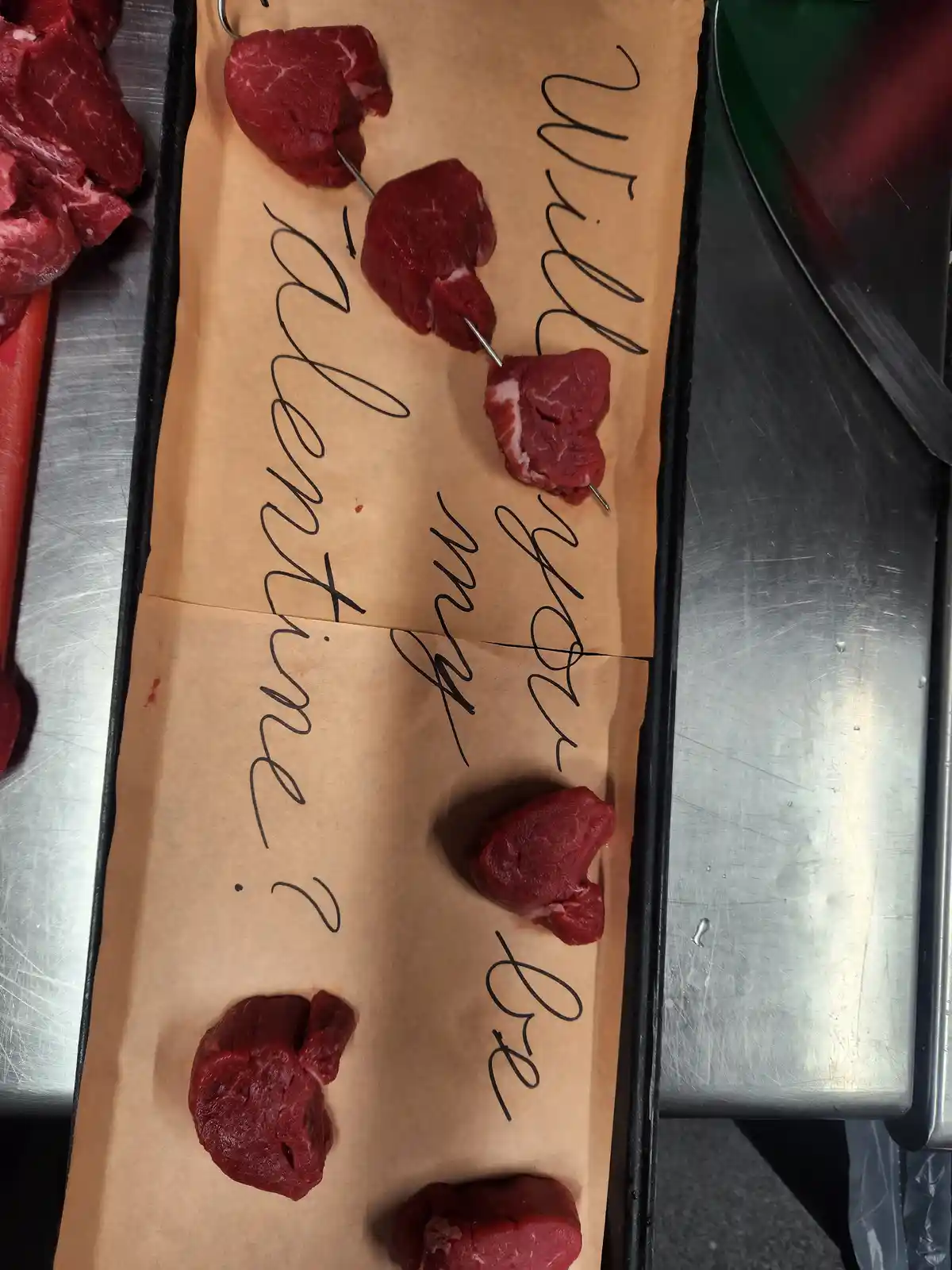 A tray lined with brown paper holds several raw meat chunks, some pierced with metal skewers, arranged to spell out the…