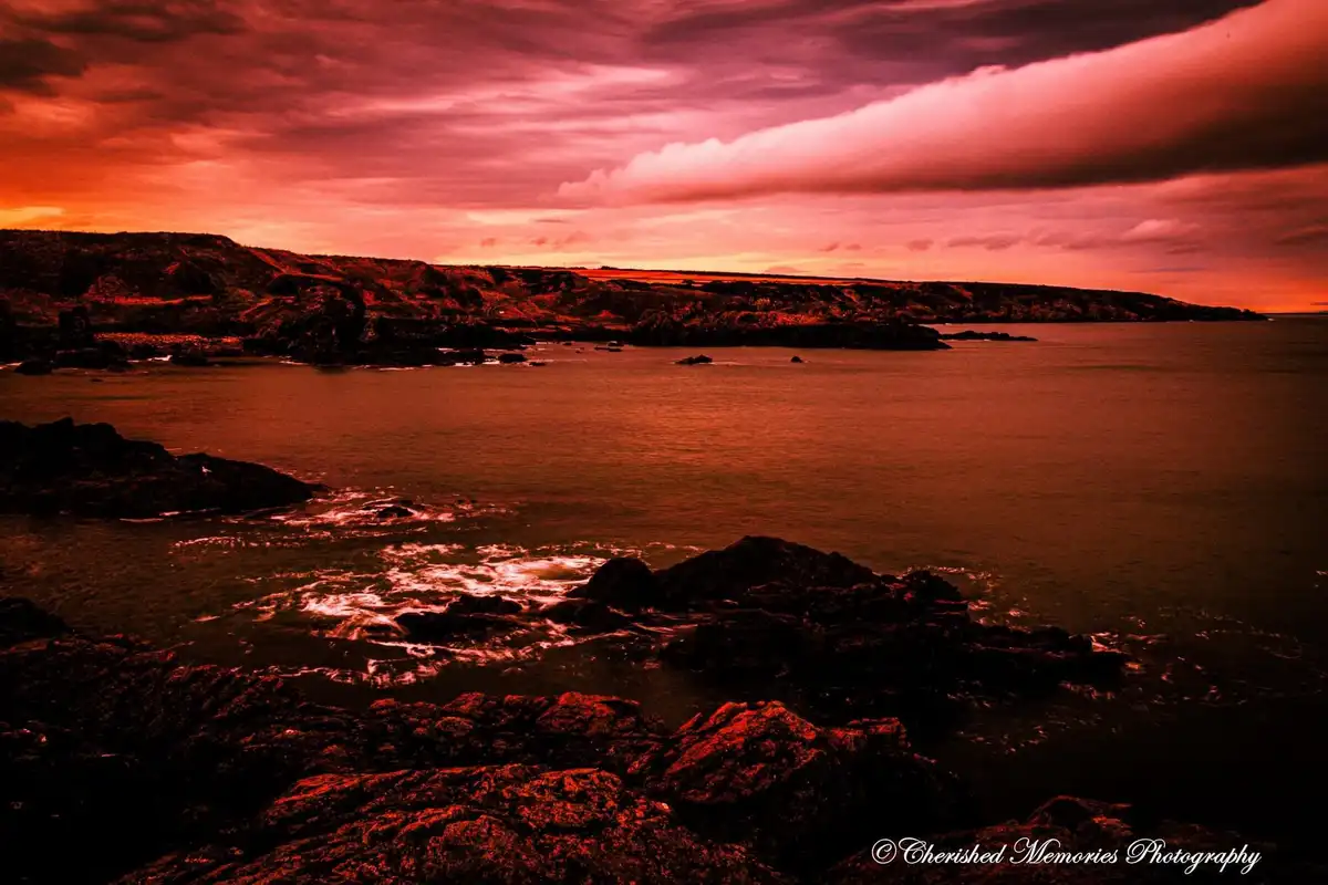 A dramatic coastal scene at sunset or sunrise, with a sky filled with fiery red and orange clouds over dark, silhouetted…