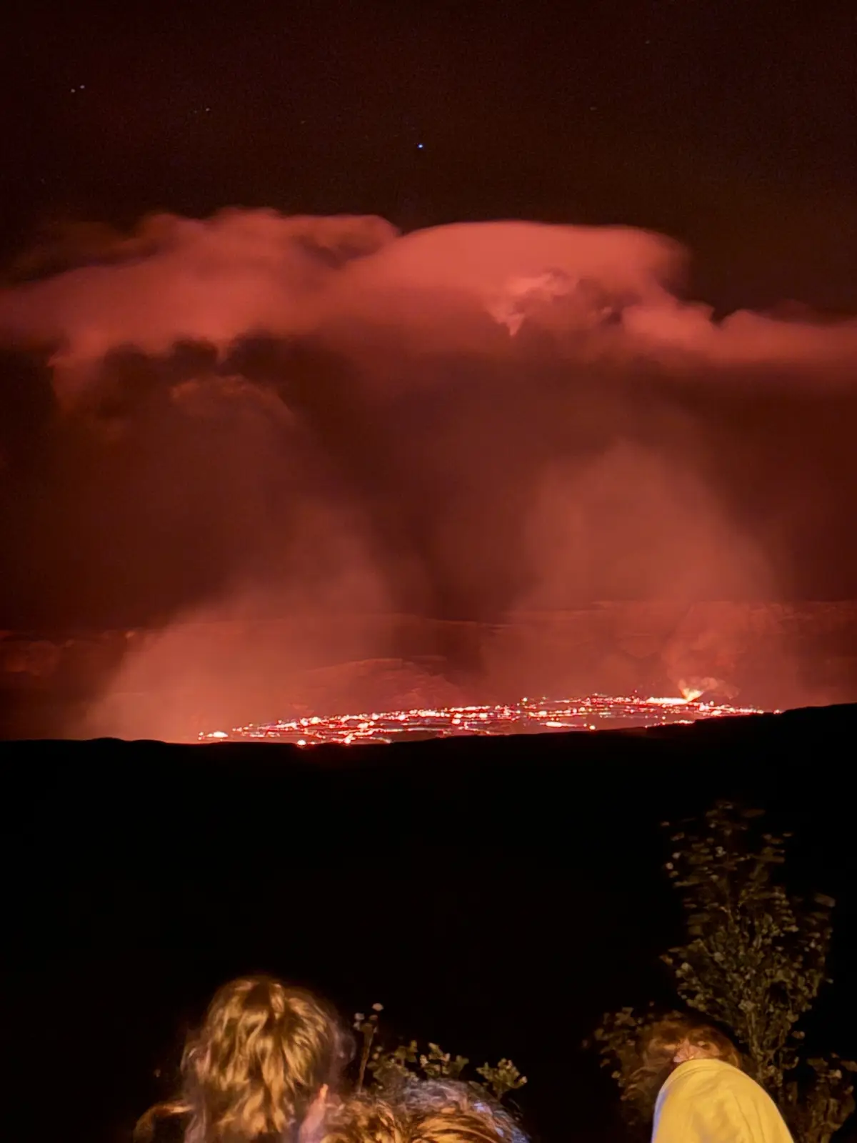 A nighttime photo shows several people from behind, looking out at a dramatic volcanic eruption. The sky is dark with…