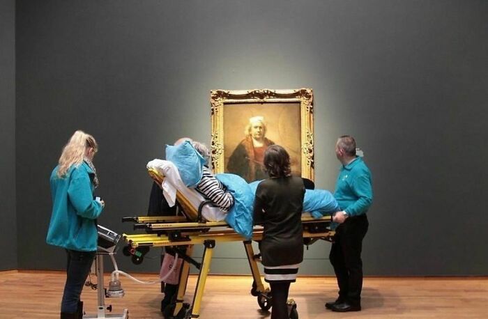 In a museum gallery, medical staff transport a patient on a gurney directly in front of a large, ornate portrait of a man…