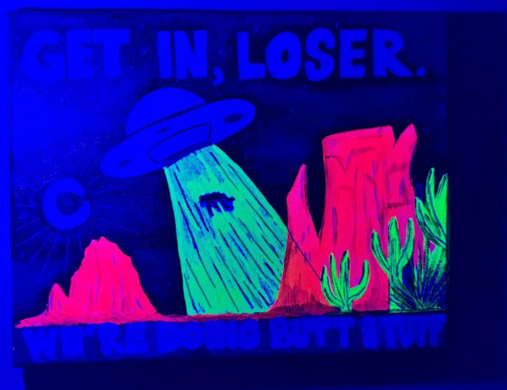 A neon-lit, UV-reactive painting depicts a UFO hovering over a desert landscape with pink rock formations and green cacti…