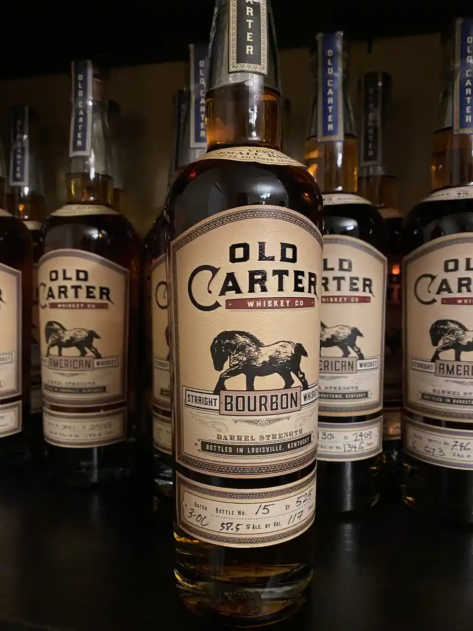 A close-up photo shows several bottles of Old Carter Straight Bourbon Whiskey lined up on a shelf, with one bottle in the…