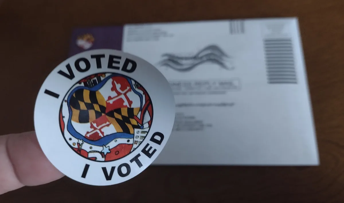 A close-up photo shows a hand holding a circular 'I VOTED' sticker featuring the Maryland state flag design. Behind it…