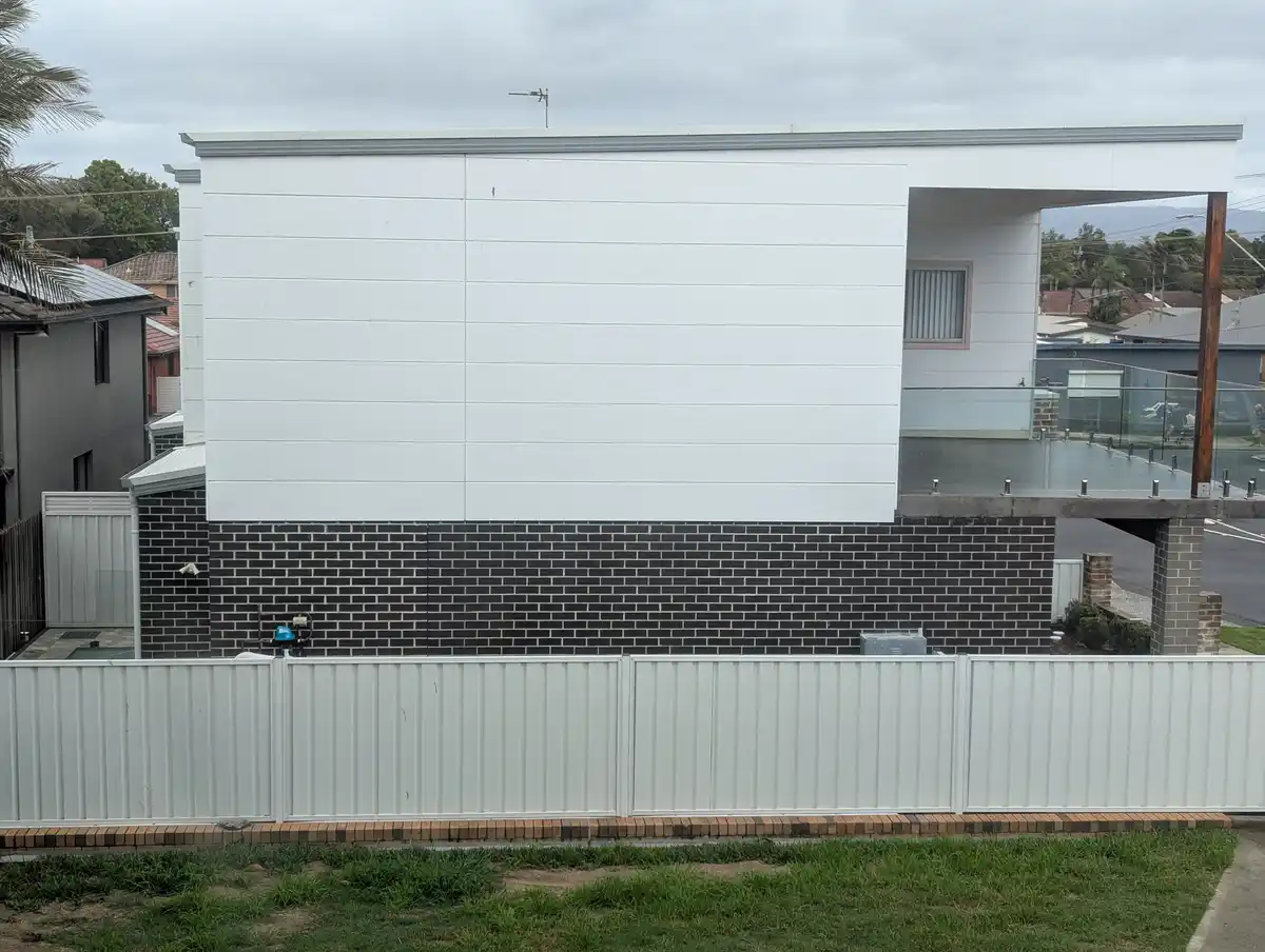 A modern house with a white upper wall and black brick lower wall, partially obscured by a white fence, with overlaid text…