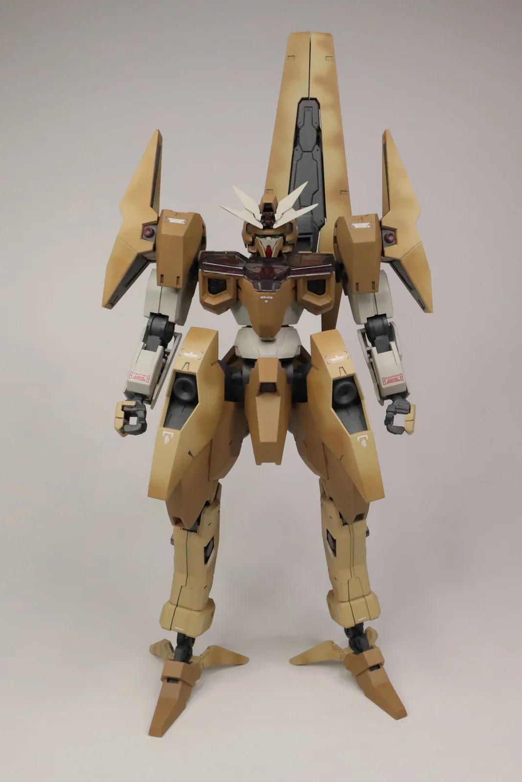 A tan and beige painted model of a Gundam-style robot stands against a plain white background. The robot has a distinctive…