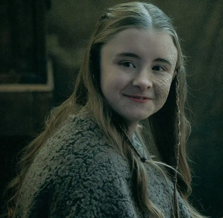 A young woman with long brown hair and braids, wearing a textured grey coat, has visible wrinkles on her face. She is…