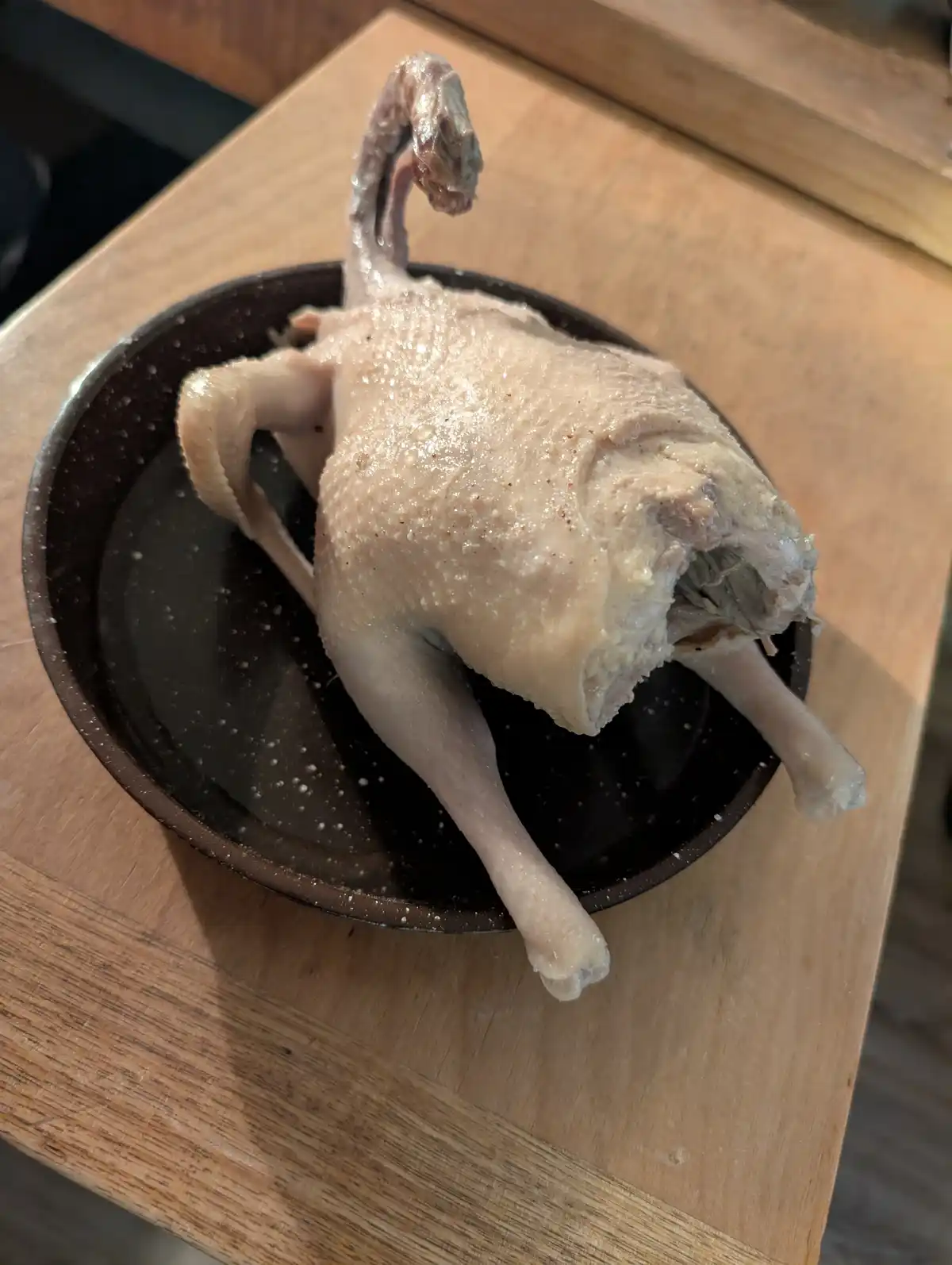 A whole raw duck, headless and plucked, sits upright in a dark bowl on a wooden surface, its neck curved upward. The…