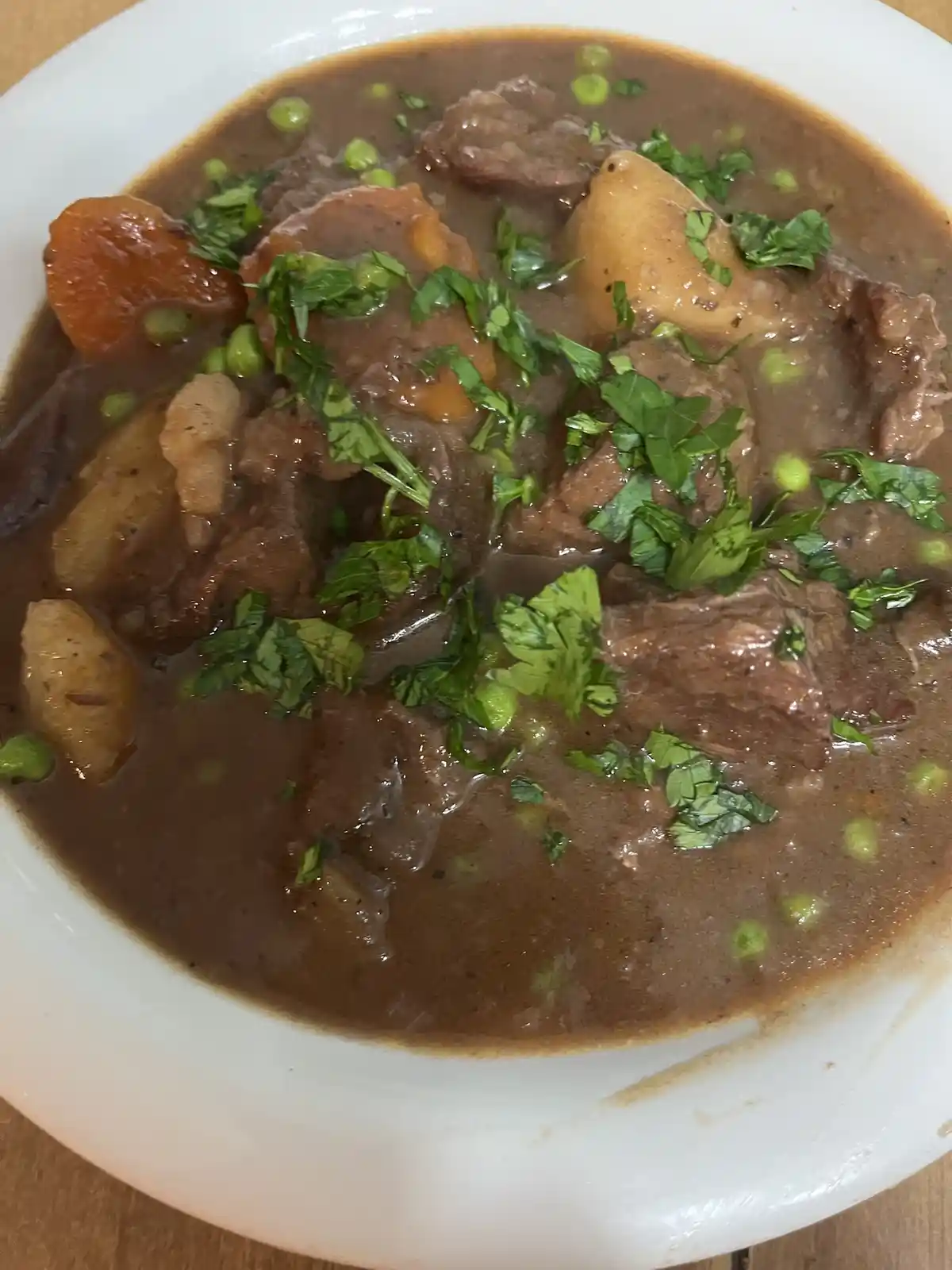 A close-up photo shows a white bowl filled with beef stew, containing chunks of meat, potatoes, carrots, green peas, and…