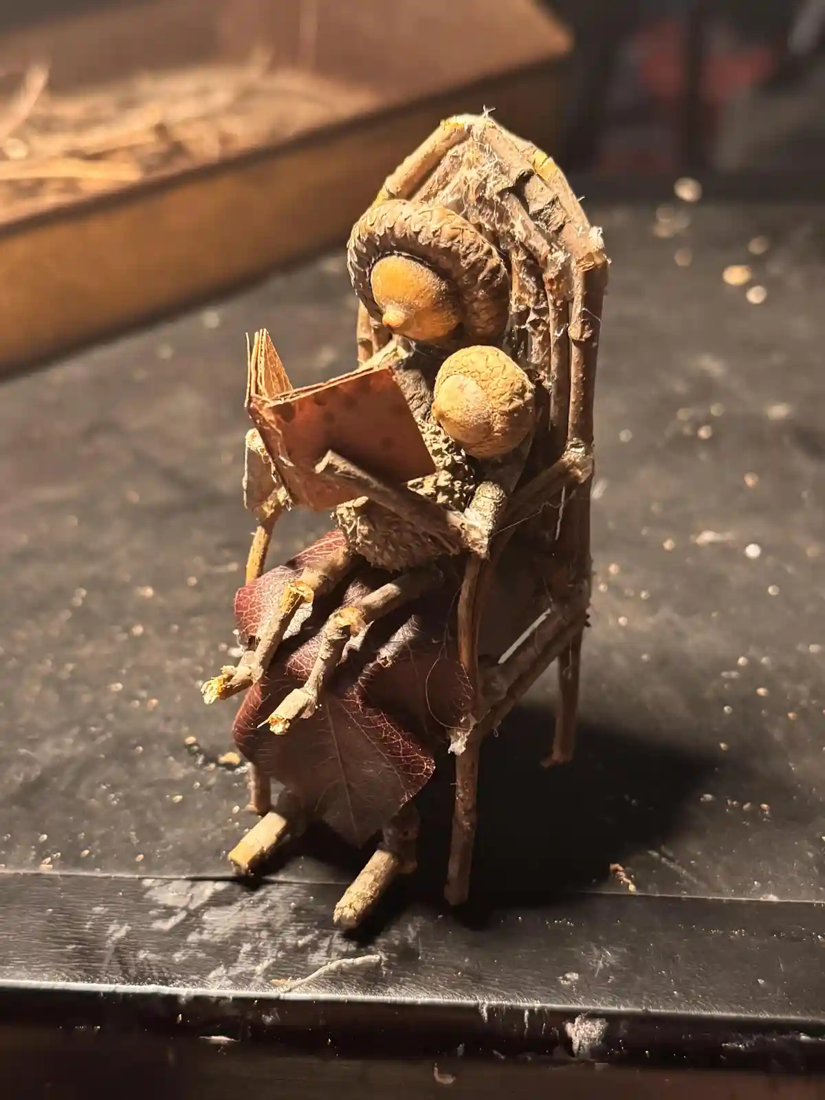 A tiny, handmade figurine crafted from natural materials like acorns, twigs, and dried leaves sits in a miniature chair…