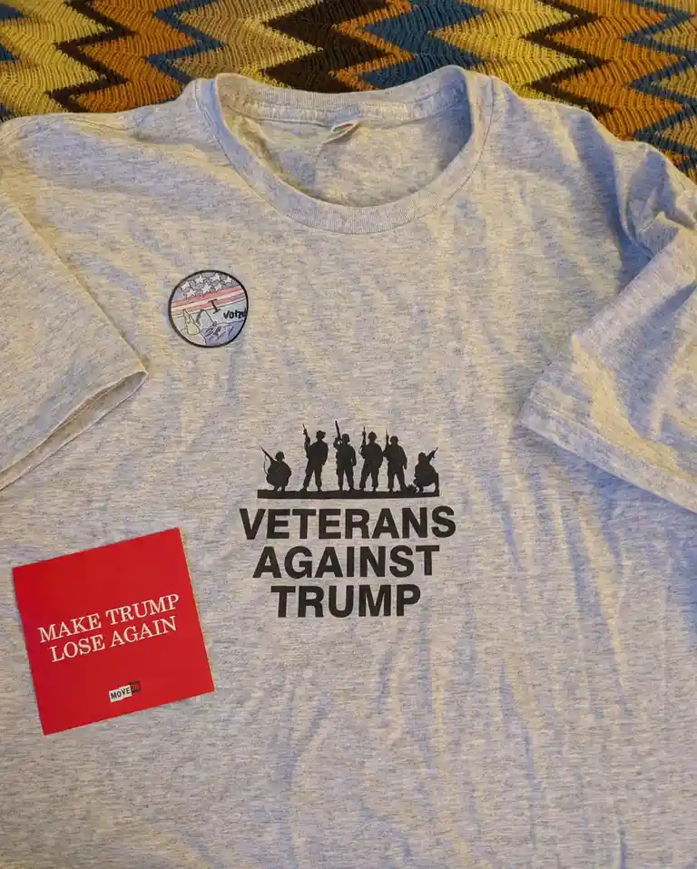 A light gray t-shirt displays a black graphic of soldier silhouettes above the text 'VETERANS AGAINST TRUMP'. A red square…