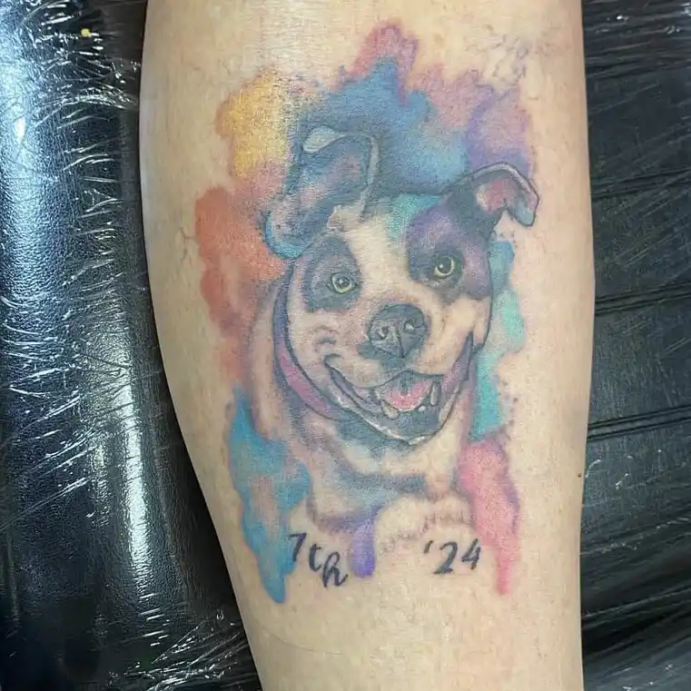 A colorful watercolor-style tattoo of a smiling dog with a collar is on a person's leg, with the text '7th '24' written…