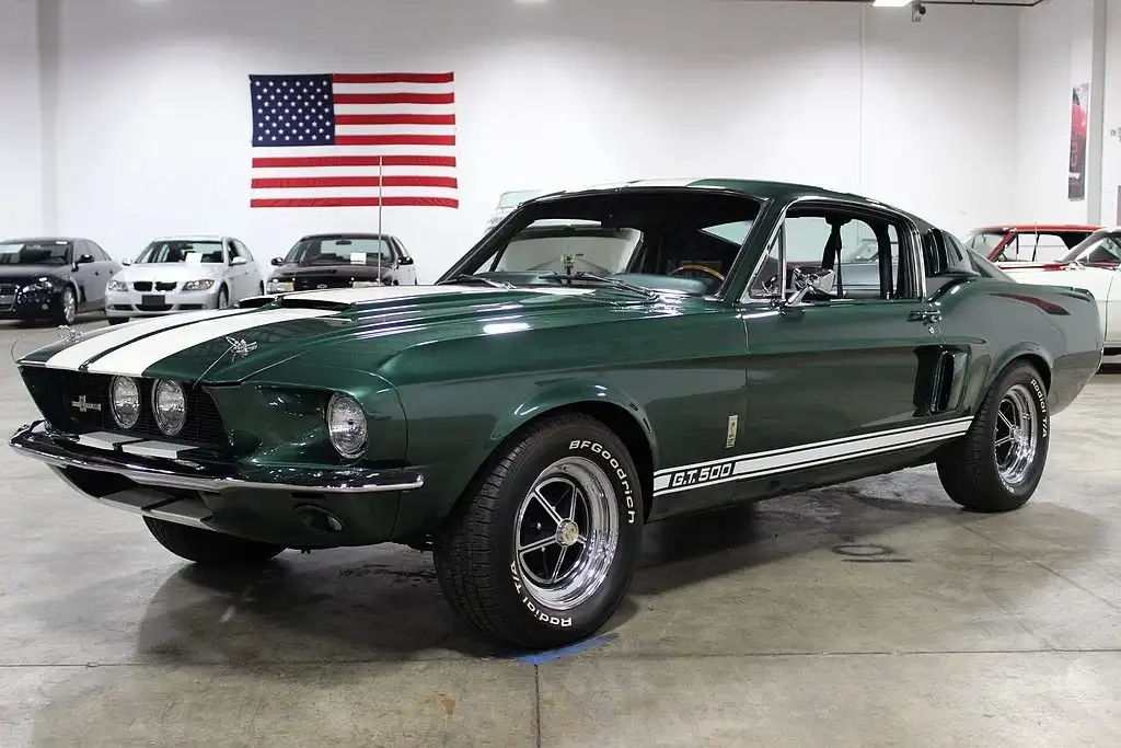 A green 1967 Ford Mustang GT500 with white racing stripes is displayed in a showroom, with an American flag hanging on the…