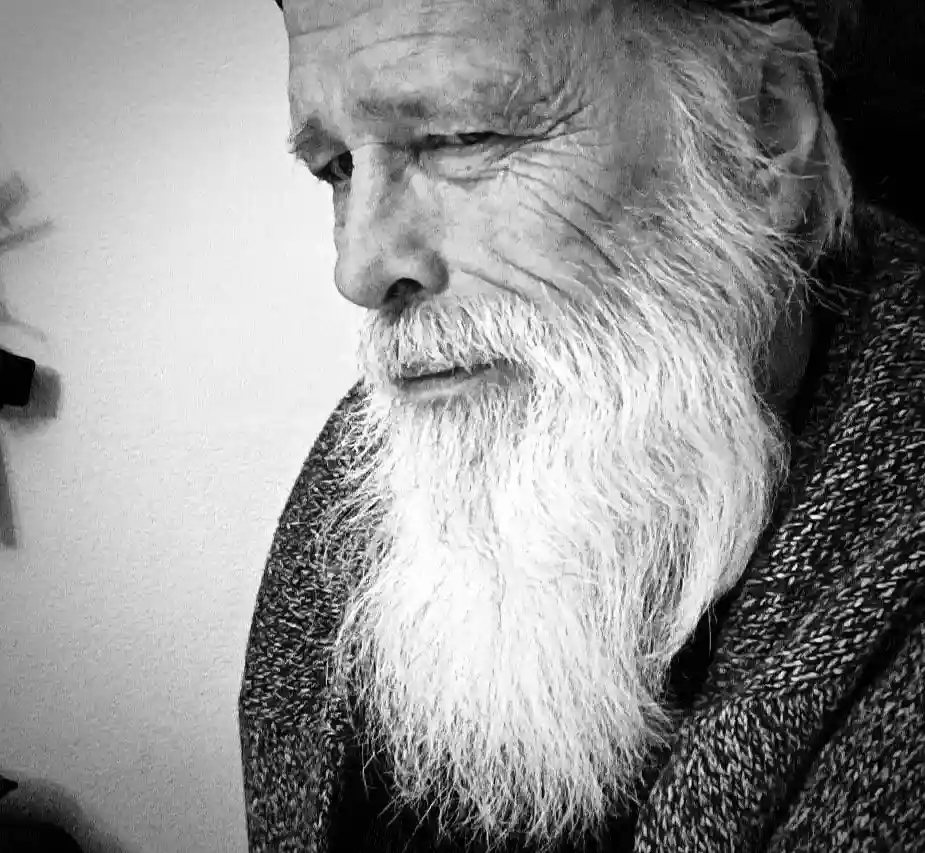 A black-and-white close-up photo of an elderly man with a long, white beard and deep wrinkles, looking downward with a…