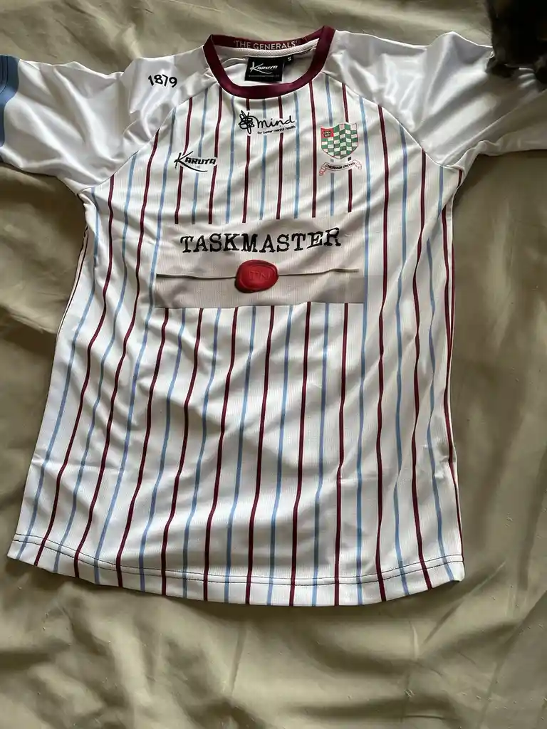 A white rugby-style jersey with red and blue vertical stripes lies flat on a beige surface. The jersey features a…