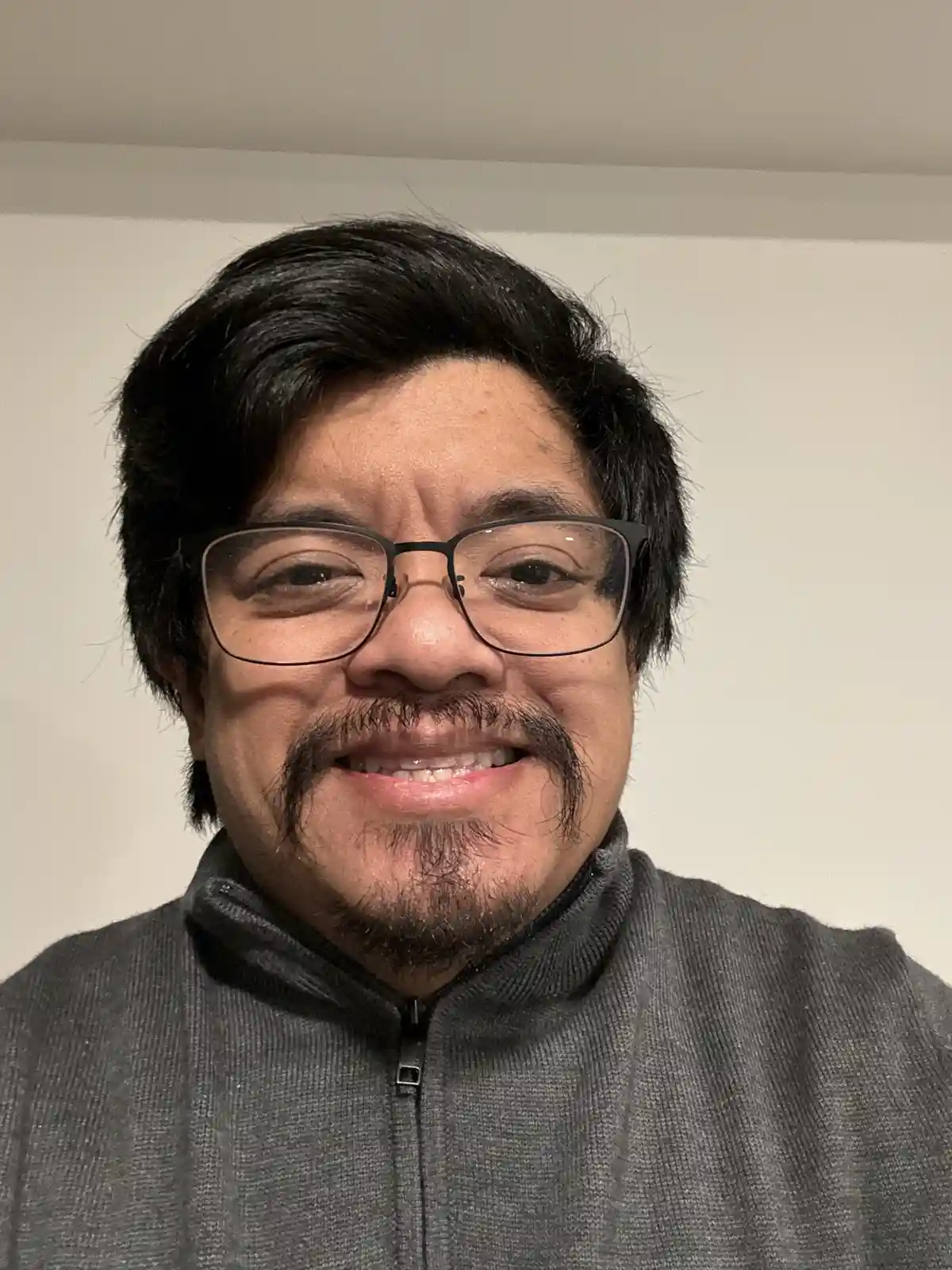 A close-up selfie of a man with dark hair, black-rimmed glasses, and a distinctive handlebar mustache, smiling while wearing…