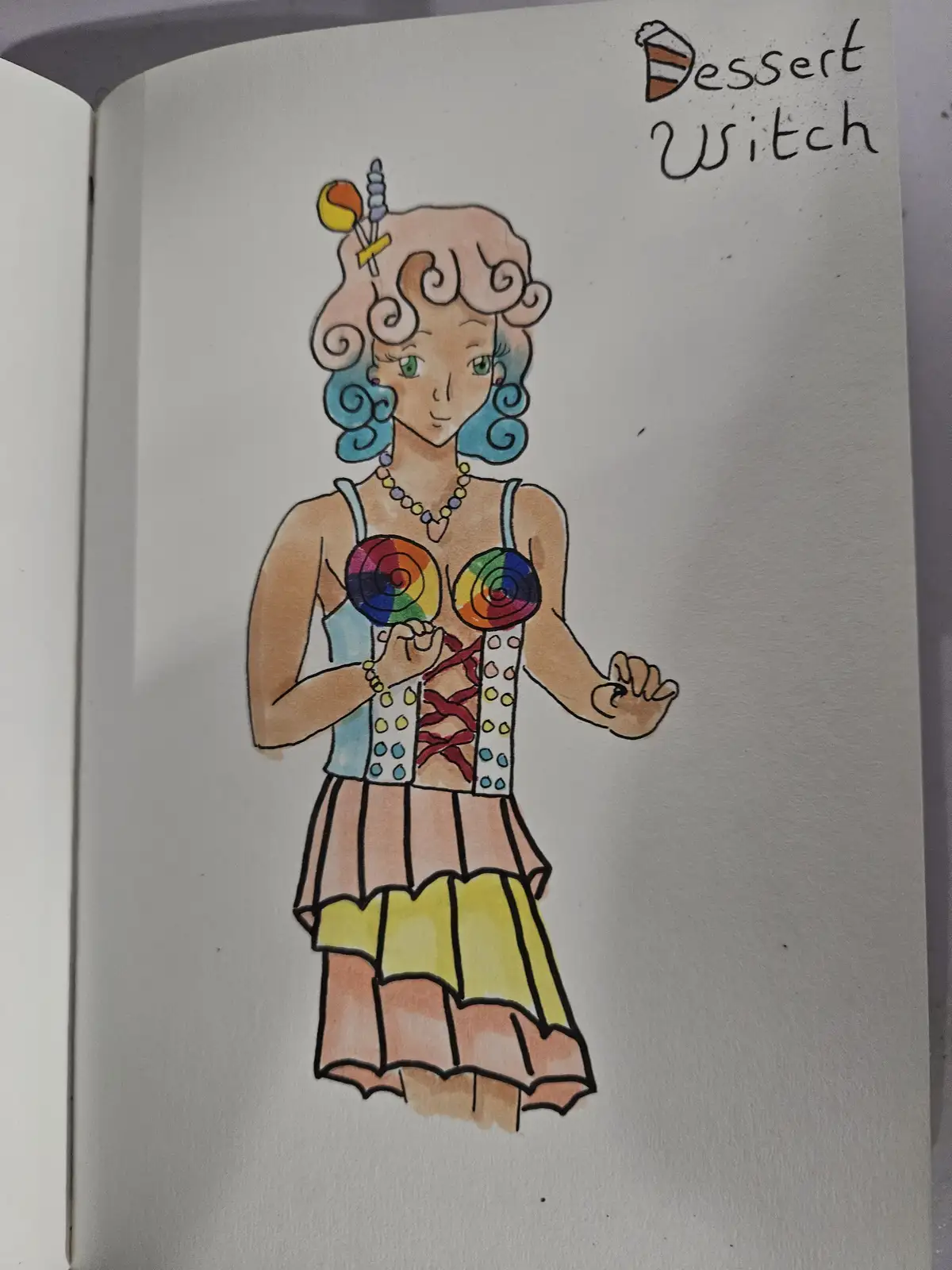 A hand-drawn illustration in a sketchbook depicts a whimsical female character labeled 'Dessert Witch'. She has curly…