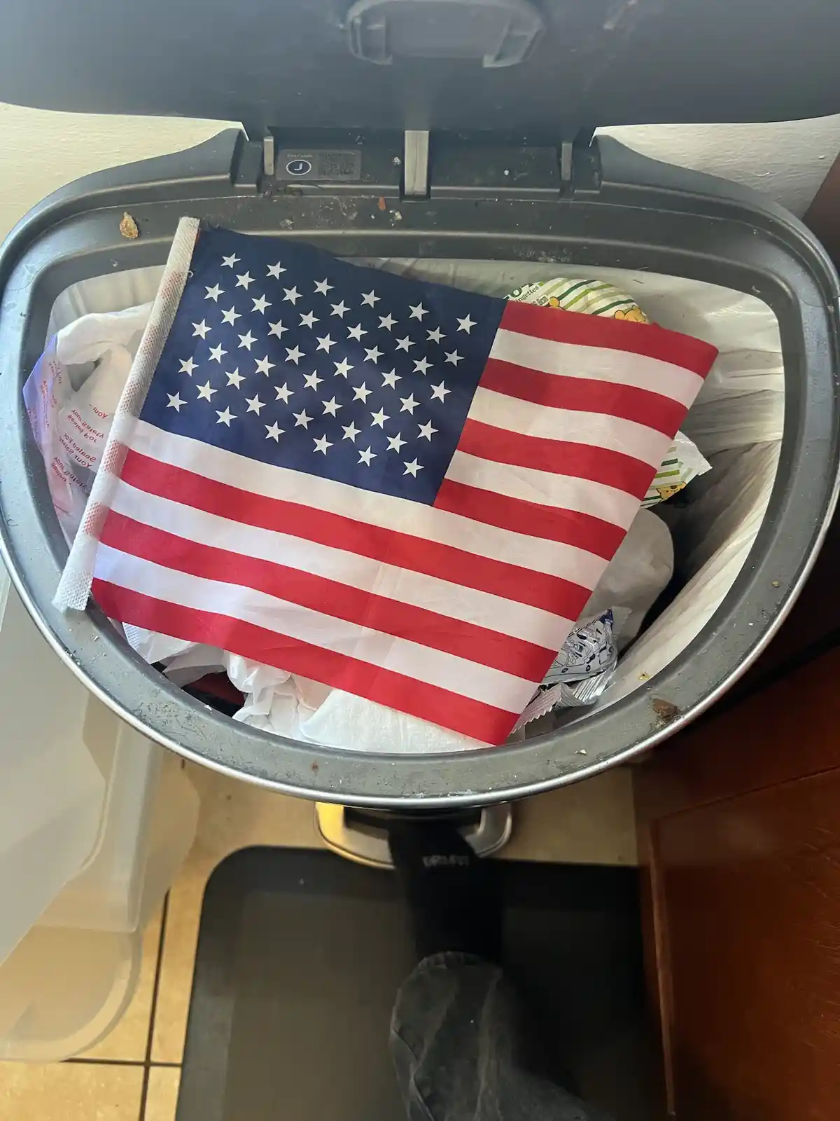 A small American flag is prominently displayed inside a household trash can, partially covering other discarded items like…