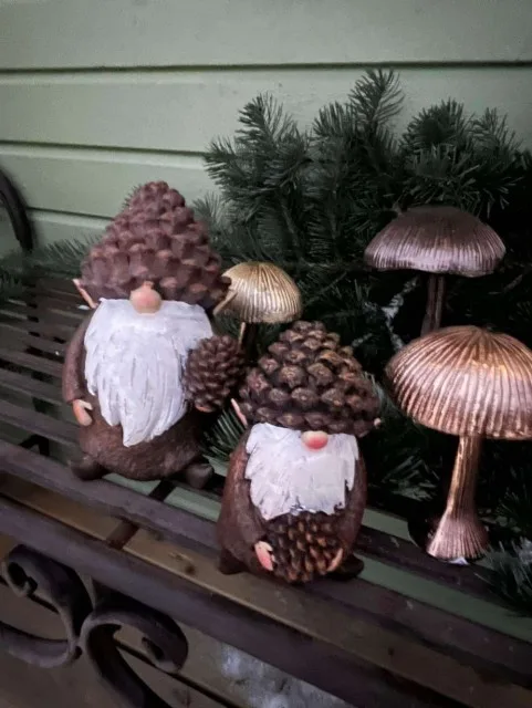 Two small decorative gnomes made from pinecones with white painted beards and pink noses sit on a metal bench beside…