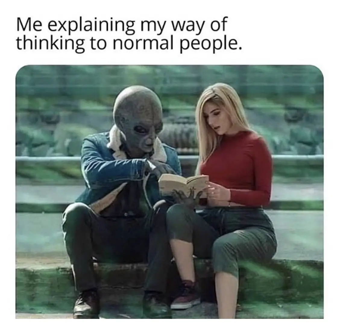 An alien with a large head and dark skin sits beside a blonde woman in a red shirt, both looking at an open book. The alien…