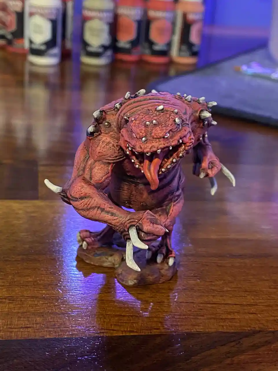 A red, spiky, monstrous figurine with sharp teeth and tusks is crouched on a wooden table, looking menacing. In the…