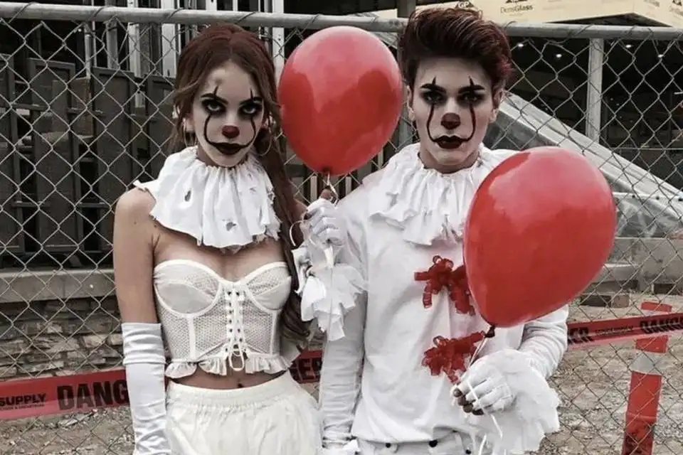 Two people dressed as clowns with white ruffled clothing, black eye makeup, red noses, and holding red balloons stand in…