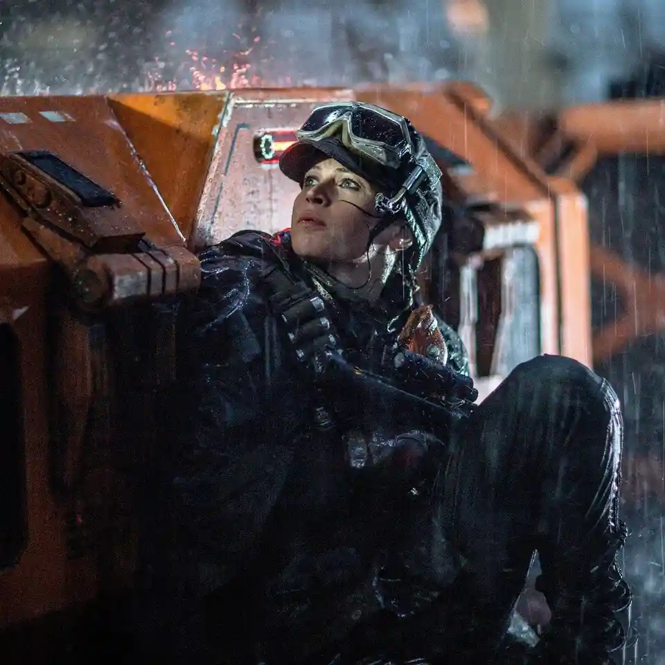 A woman in tactical gear, goggles on her head, and rain falling around her, looks upward with a determined expression while…