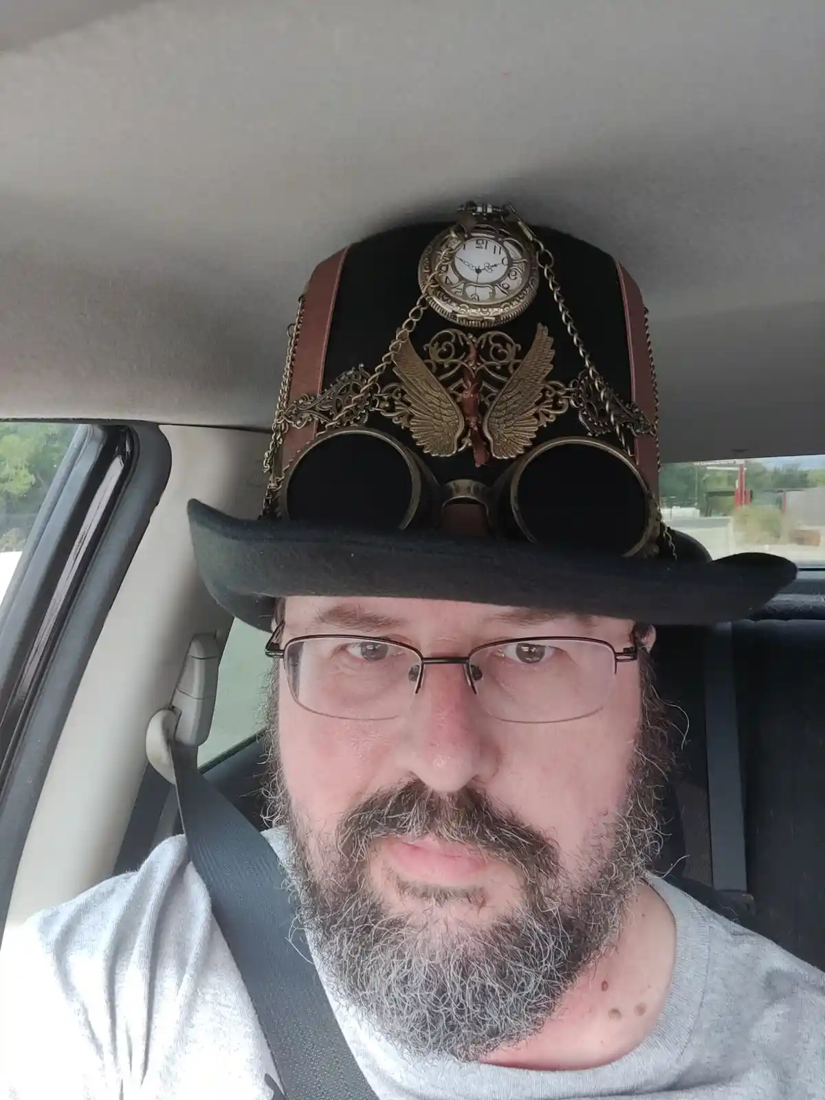 A man with glasses and a gray beard sits in a car wearing an elaborate steampunk hat adorned with a pocket watch, chains…